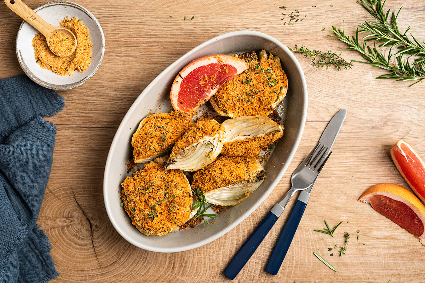 Gebackener Fenchel in Auflaufform mit Grapefruit-Schnitzen und Rosmarin-Zweig auf einem Holztisch